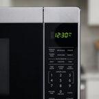 A macro close-up image showing a stainless steel microwave control panel with a digital clock and various buttons.
